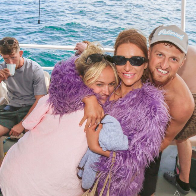 Three friends smiling and hugging on a boat; one wears a purple fuzzy jacket and sunglasses, another is in pink, and a shirtless man in a cap leans in. Ocean water is visible in the background.