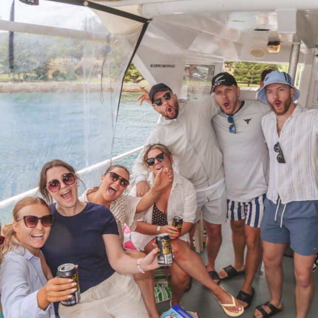 A group of smiling people wearing sunglasses and summer clothes sit on a boat, holding drinks and posing energetically for the camera, with water and trees visible through the windows.