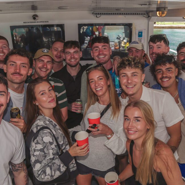 A group of young adults, both men and women, smiling and holding drinks while posing together on a boat. The background shows windows with views of water and part of a harbor. The mood is cheerful and festive.