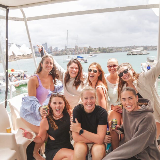 A group of eight smiling friends pose together on a boat with city buildings, the Sydney Opera House, and other boats visible in the background under a cloudy sky.