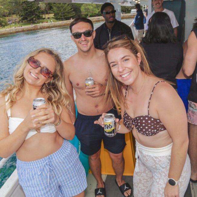 A group of people in swimwear enjoying drinks on a boat, smiling at the camera. The background shows blue water, trees, and clear skies, suggesting a lively and sunny outdoor gathering.