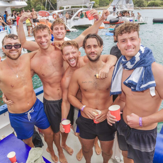 Six young men in swim trunks stand on a boat, smiling and posing for a photo. Some have their arms around each other and hold red cups. There are boats, water, and other people in the background, suggesting a lively party atmosphere.