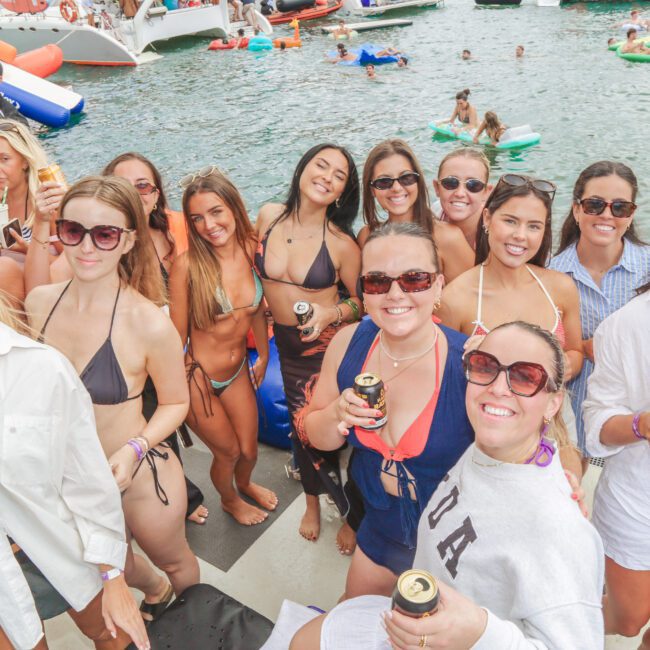 A group of smiling people in swimwear and sunglasses gather on a dock, holding drinks, with boats and others swimming in the background on a sunny day.