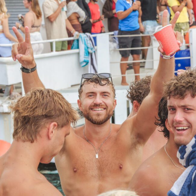 A group of young men in swimwear on a boat, one smiling and raising his arms in celebration while holding a red cup. Other people and a lifeboat are visible in the background, suggesting a lively party atmosphere.