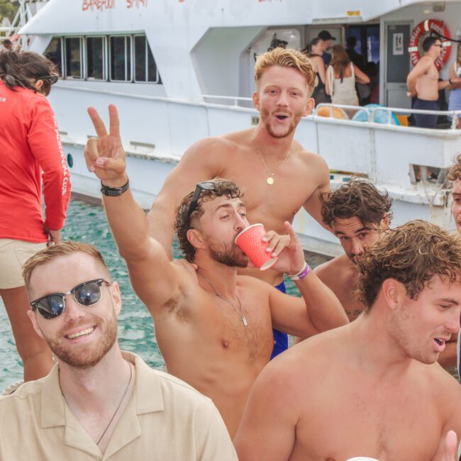 A group of young adults in swimwear enjoy a lively boat party. One man makes a peace sign, another holds a red cup, and others smile at the camera. The boat and water are visible in the background.