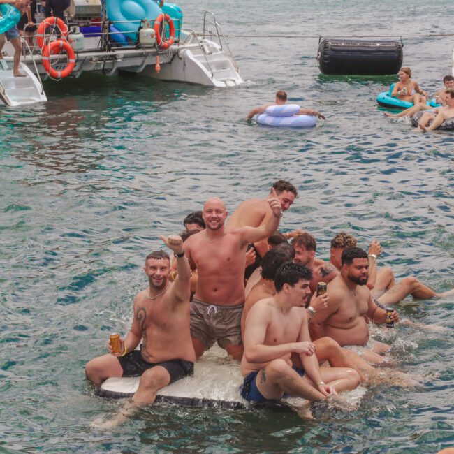 A group of men in swimwear happily pose on a floating platform in the water while others swim and relax nearby. Boats and more people are visible in the background, creating a lively, party atmosphere.