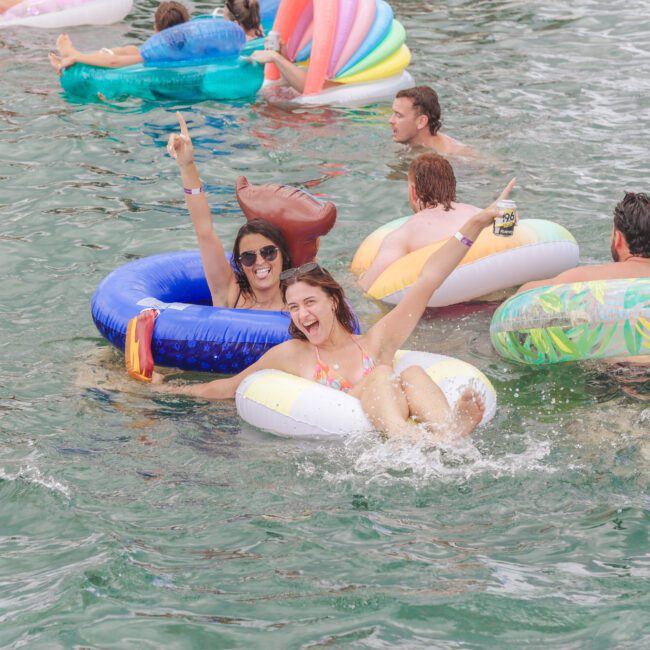 Two women in colorful inflatable rings smile and raise their arms while floating on water. Other people on floaties are nearby, enjoying a lively, fun atmosphere. The scene captures summer relaxation and celebration.