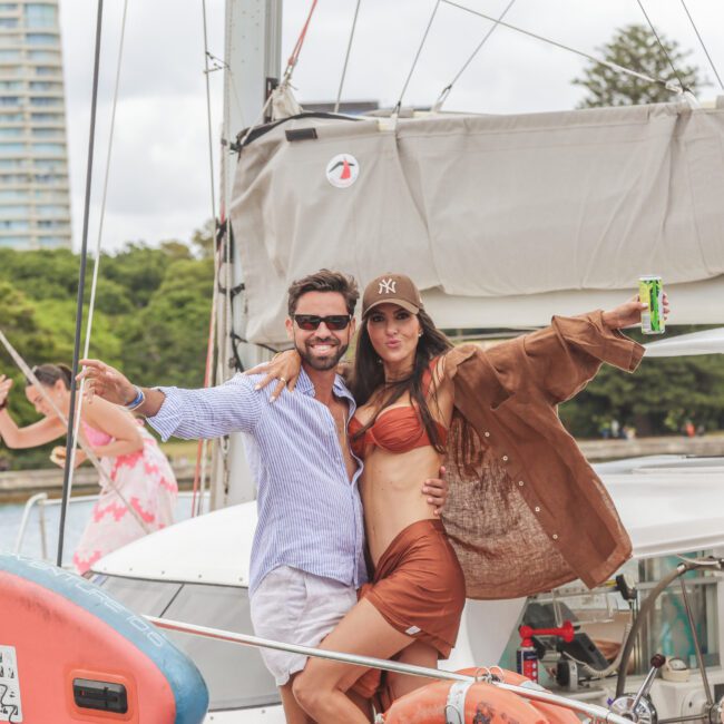A smiling man and woman pose together on a boat. The woman wears a brown bikini and matching shirt, with a cap, holding a can. Other people relax on the boat, with buildings and trees in the background.