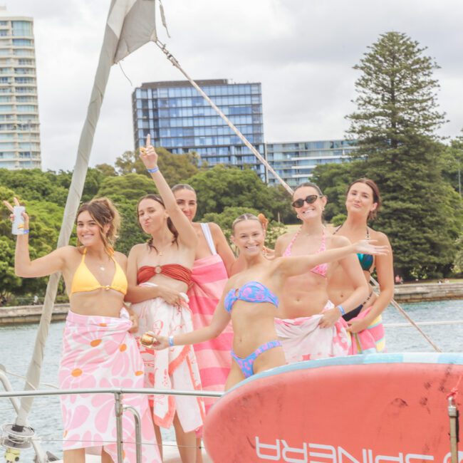 Six smiling women in swimsuits and towels stand on a sailboat, holding drinks and posing cheerfully. A paddleboard is in the foreground, with trees and tall buildings visible in the background by the water.