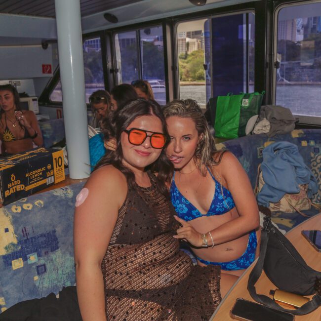 Two women in swimwear pose and smile on a boat's lounge area with other people relaxing in the background. There are bags, drinks, and snacks on the table, and city buildings visible through the windows.