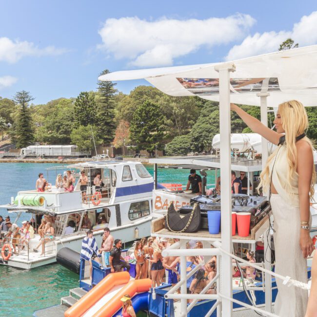 A group of people party on boats docked near a shoreline with trees. Some are on the deck, others in the water. A woman in the foreground takes a selfie, and the sky is blue with scattered clouds.