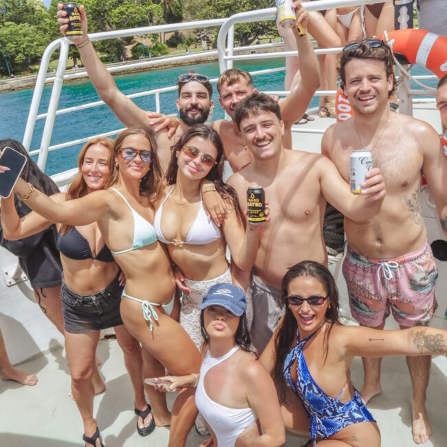 A group of young adults in swimsuits smile and raise drinks while posing together on a boat. The background shows bright blue water and green trees, suggesting a sunny day on a lake or at sea.
