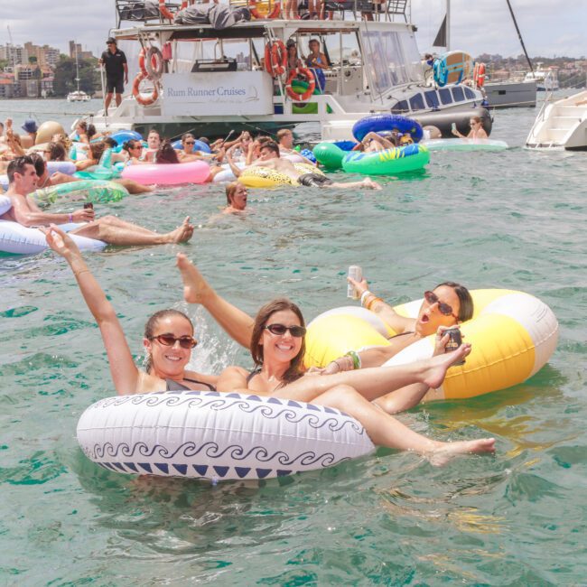 A group of people on colorful inflatable rings relax and smile in the water near several boats during a lively summer party. Three women in the foreground pose and wave at the camera.