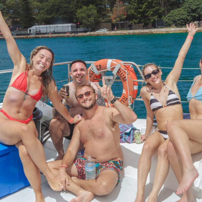 Five people in swimsuits sit on a boat deck, smiling and raising their arms in celebration. The boat is on blue water near a forested shore under a sunny sky. There is a life preserver and a blue cooler nearby.