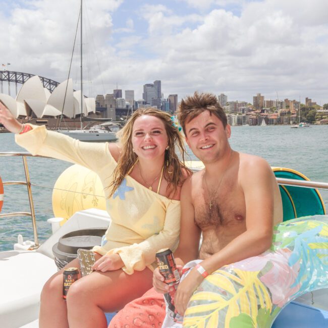 A smiling woman and man sit on a boat holding drinks, with the Sydney Opera House and Harbour Bridge in the background. They appear to be enjoying a sunny day, with inflatable pool floats nearby.