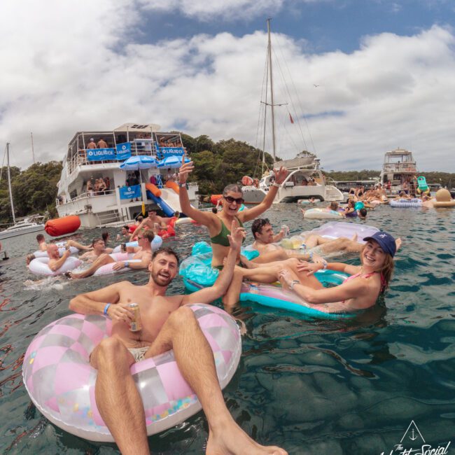 A group of young adults relax and laugh on colorful pool floats in the water near boats, enjoying a sunny day. Other people float nearby, with yachts and trees in the background. The atmosphere is lively and festive.