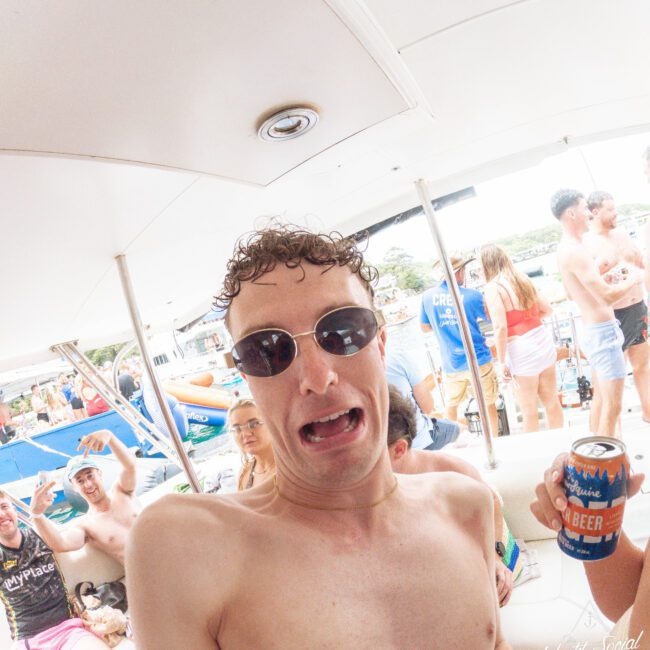 A shirtless man with curly hair and sunglasses makes a silly face while holding a can of beer on a boat. Other people in swimwear are socializing in the background.