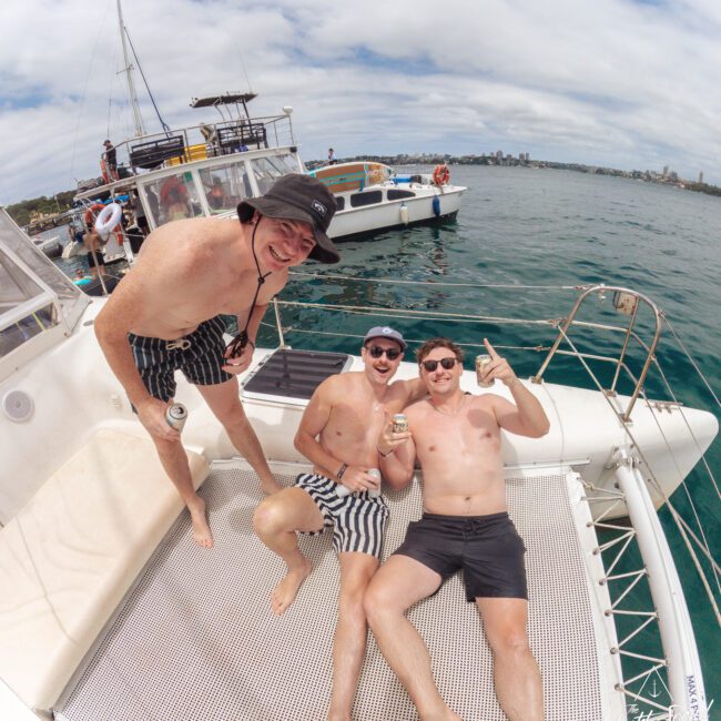 Three smiling men in swim trunks relax on a boat, with two lying down and one standing, holding drinks. Other boats and a city skyline are visible in the background under a partly cloudy sky.