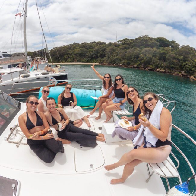 Eight women in swimsuits relax on a boat, smiling and enjoying drinks and snacks. There are trees and water in the background, with another boat nearby under a partly cloudy sky.
