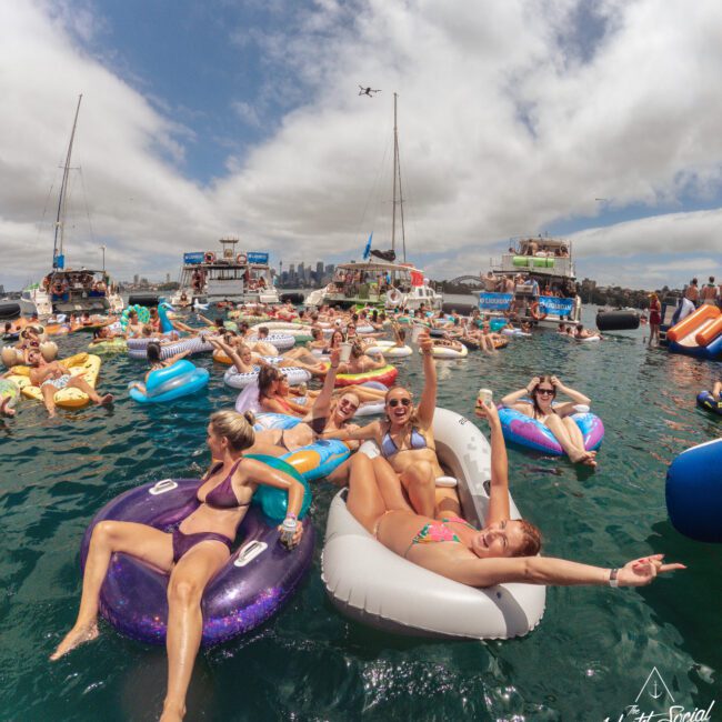 A group of people relax on inflatable rafts in the water, celebrating and posing for photos near anchored boats. The sky is partly cloudy, and the city skyline is visible in the background.