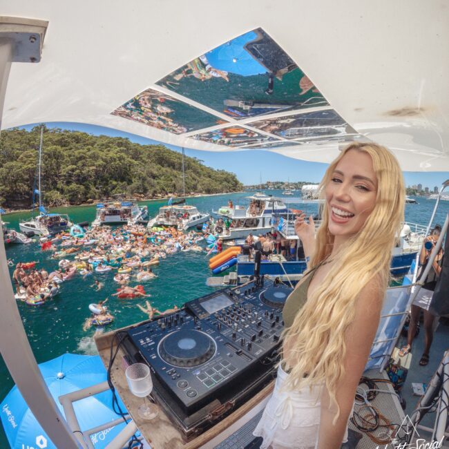 A smiling woman with long blonde hair stands at a DJ booth on a boat, overlooking a lively crowd of people swimming and gathering on boats in a blue-green body of water, surrounded by trees and sunshine.