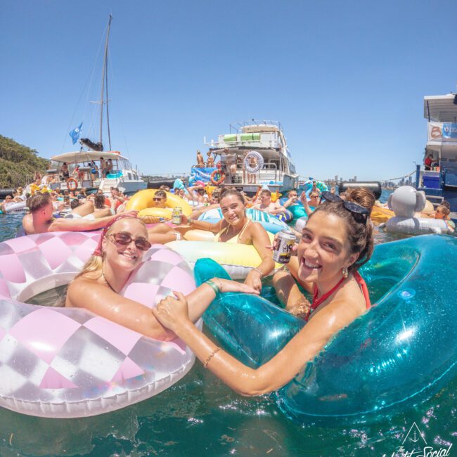 Three smiling women in swimsuits hold hands while floating on inflatable pool rings in the ocean, surrounded by other people on floaties and boats, under a clear blue sky.