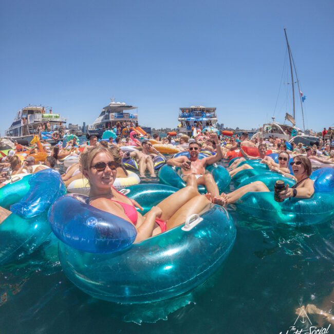 A large group of people relax on inflatable tubes in the water near several boats under a clear, sunny sky. Many are smiling, wearing swimsuits and sunglasses, enjoying a festive, social gathering.