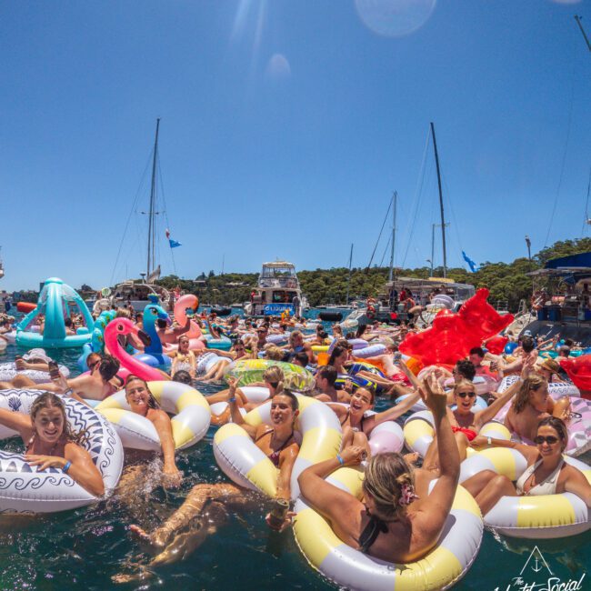 A group of people relaxing and smiling on colorful inflatable pool floats in the water, surrounded by yachts under a clear blue sky at a lively outdoor event.