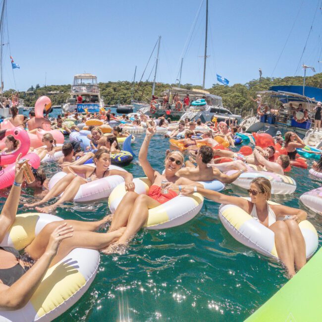 People float on colorful inflatable rings in a crowded party on the water near anchored yachts, under a clear blue sky. Everyone looks relaxed and festive, enjoying the sunny day.