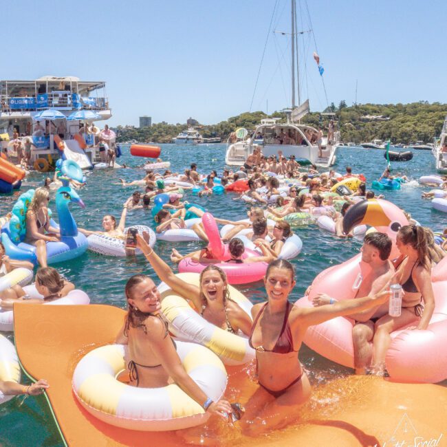 A lively group of people in swimsuits float on colorful inflatables at a sunny yacht party, with boats and clear blue water in the background. Everyone is smiling and enjoying the festive summer atmosphere.