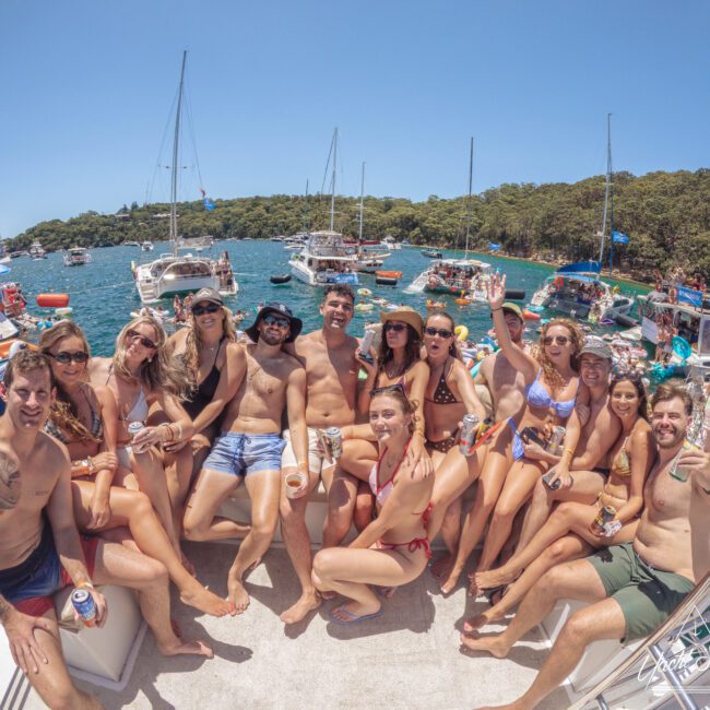 A group of people in swimwear sit closely together, smiling and posing on a boat surrounded by water and anchored yachts, with trees visible in the background under a clear blue sky.