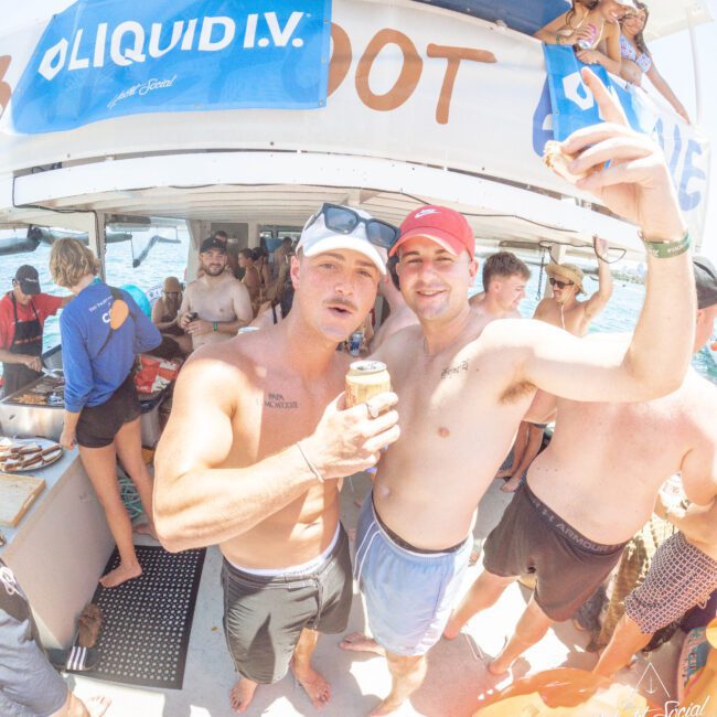 Two men in swim trunks and baseball caps stand on a sunny, crowded boat, smiling and holding cans. Other people swim, relax, and socialize around them. A “Liquid I.V.” banner hangs from the upper deck.