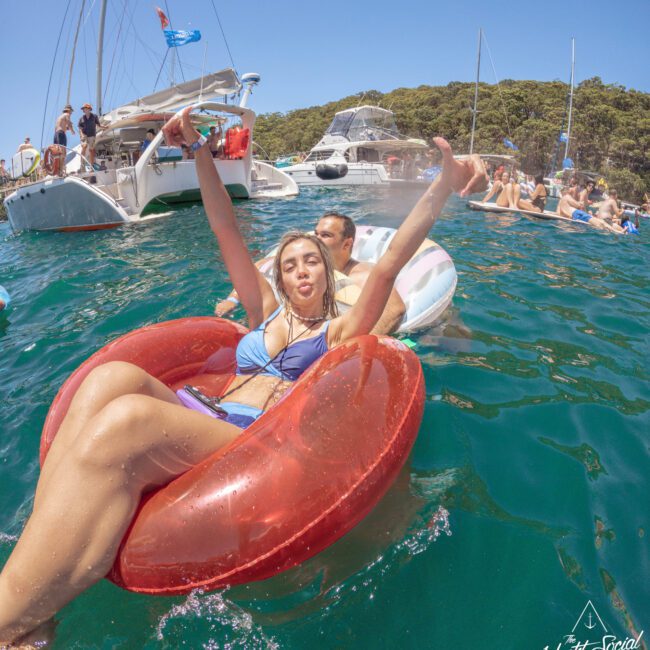 A young woman in a blue bikini sits on a red inflatable ring, smiling and making a peace sign in the water near yachts. Other people relax on inflatables and swim in the background under a clear, sunny sky.