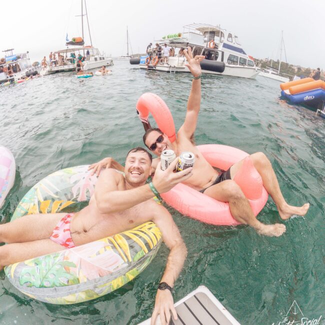 Two men in swimwear float on inflatable pool tubes in the water, smiling and raising beer cans. Boats and people are in the background, suggesting a lively party atmosphere on the water.