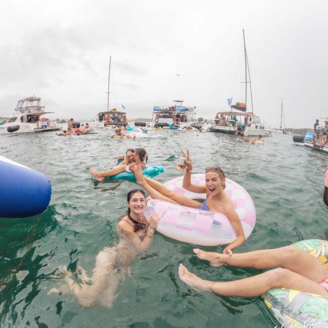 People are enjoying a party on the water, surrounded by boats. Two smiling women are in the foreground, one sitting on a pink float and flashing a peace sign while the other swims beside her. The atmosphere is festive and relaxed.