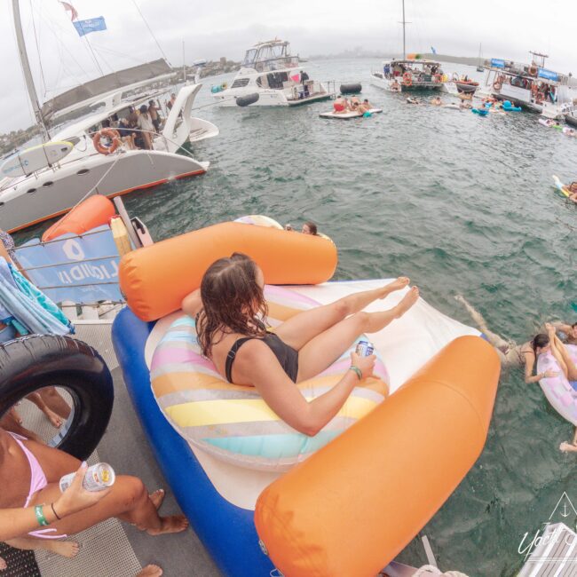 A woman lounges on a colorful inflatable chair on a boat, holding a drink. Around her, people swim and float in the water near several anchored boats during a lively summer party.