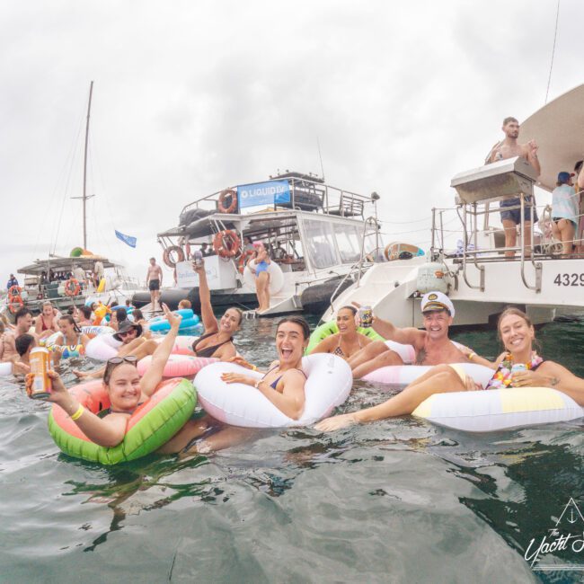 A group of people in colorful pool floats enjoy themselves in the water near anchored boats. Some hold drinks and smile at the camera, while others stand on the boats. The atmosphere is lively and festive.