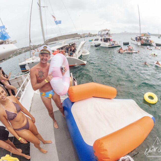 People on a boat enjoy a party with inflatables and swimwear; a man in patterned swim trunks holds a donut float and stands by an inflatable slide, with other boats and swimmers in the water nearby.