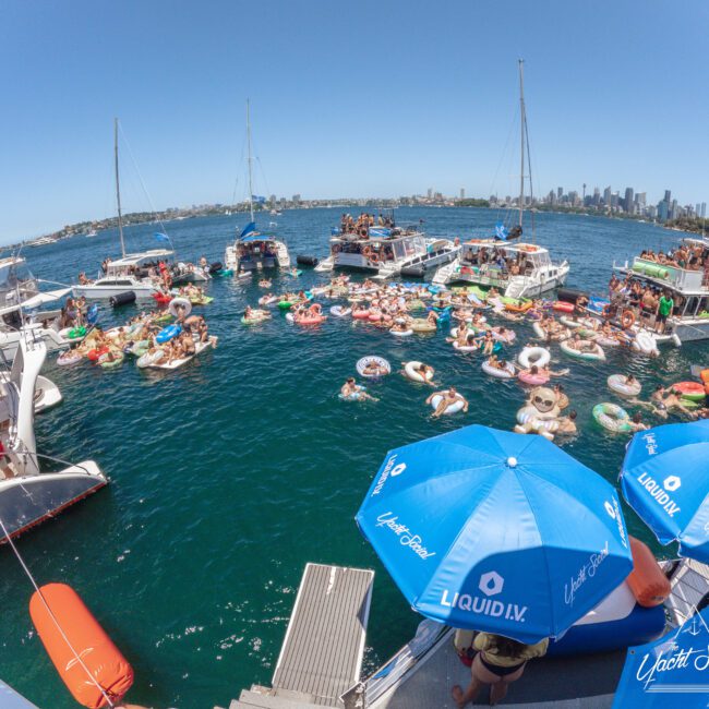 A large group of people enjoy a party on boats and colorful floaties in the water, with city skyscrapers visible in the distant background under a clear blue sky. Blue umbrellas with "Liquid I.V." logo are in the foreground.