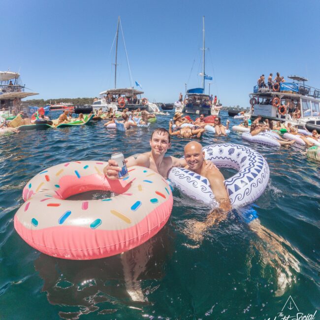 Two men in inflatable pool floats, one shaped like a donut, smile in the water holding drinks. Several people relax on other floats and boats nearby under a clear blue sky. Logo in corner reads "Yacht Social Club.