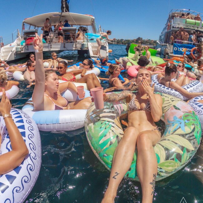 A group of people lounge on colorful inflatable tubes in the water near boats, enjoying drinks and socializing under the sun at a lively outdoor party. The scene is vibrant and festive.