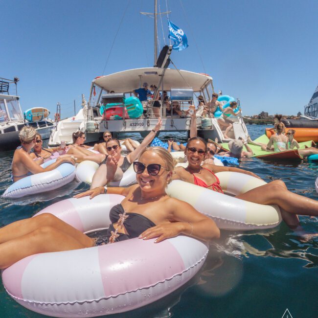 A group of people relax in inflatable pool rings on the water near a yacht, smiling and enjoying a sunny day at a lively boat party. Other yachts and blue sky are visible in the background.