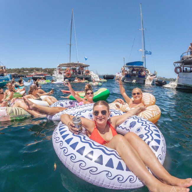 A group of smiling people relax on inflatable pool floats in the sea, holding drinks, with several boats anchored nearby under a clear blue sky.