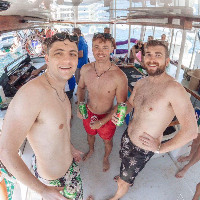 Three men in swim trunks holding drinks smile at the camera on a boat. Other people are in the background, some seated and some standing. The setting appears relaxed and festive, with sunlight streaming in.