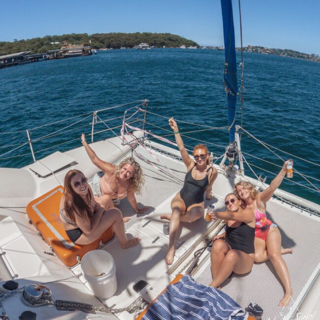 Five women in swimsuits relax and smile on the deck of a sailboat, raising their drinks and enjoying a sunny day on the water, with scenic shoreline and trees visible in the background.