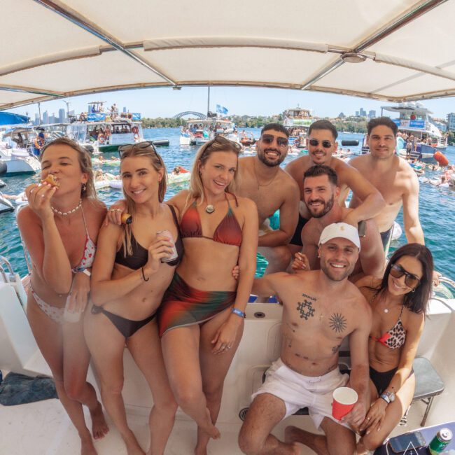 A group of nine smiling adults in swimsuits pose on a boat with a crowded lake, boats, and city skyline in the background on a sunny day. Some hold drinks, and the atmosphere is festive and relaxed.