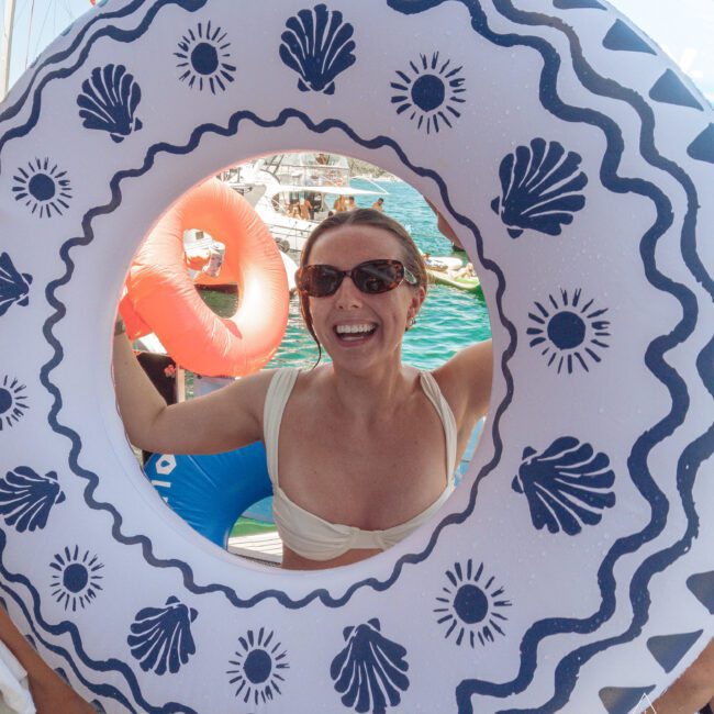 A smiling woman wearing sunglasses poses through the center of a large, white inflatable swim ring decorated with blue seashell patterns. Boats and water are visible in the sunny background.