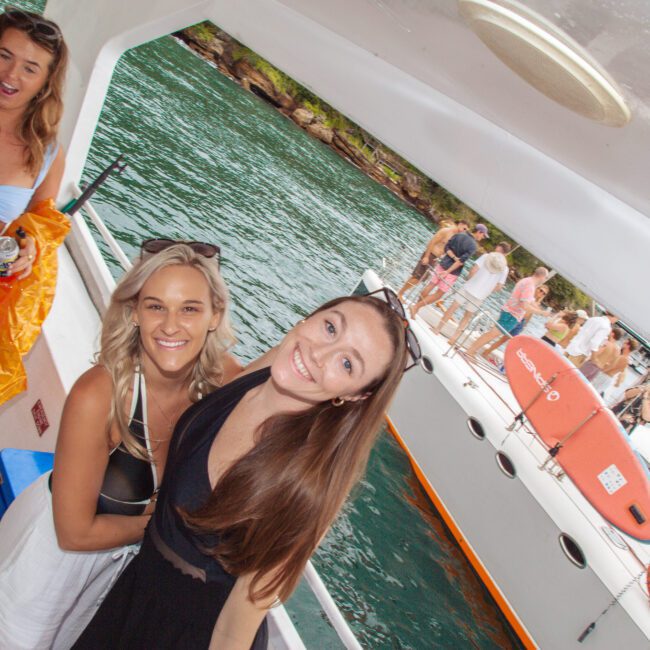 A group of young women smile and pose on a boat, with water and another boat full of people in the background. The atmosphere appears lively and festive, suggesting a fun gathering or party on the water.