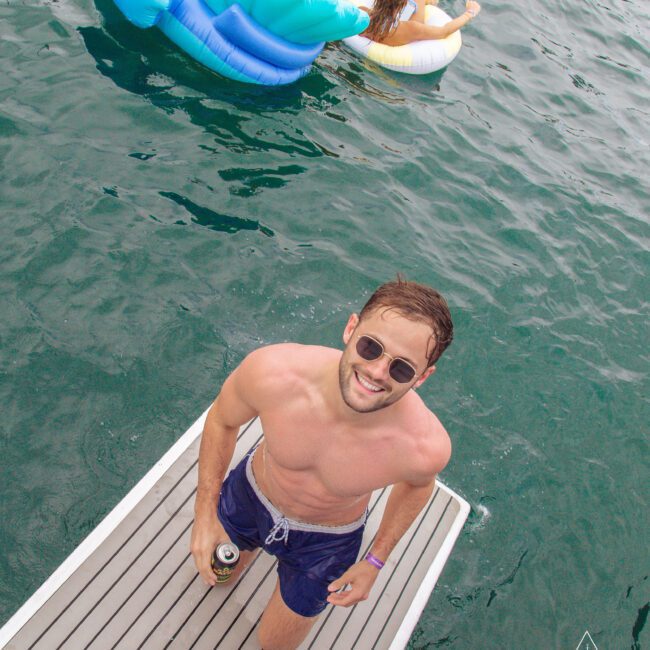 A man in sunglasses stands on a dock holding a drink, smiling up at the camera. In the background, two people relax on inflatable floats in the water. The scene appears fun and summery.