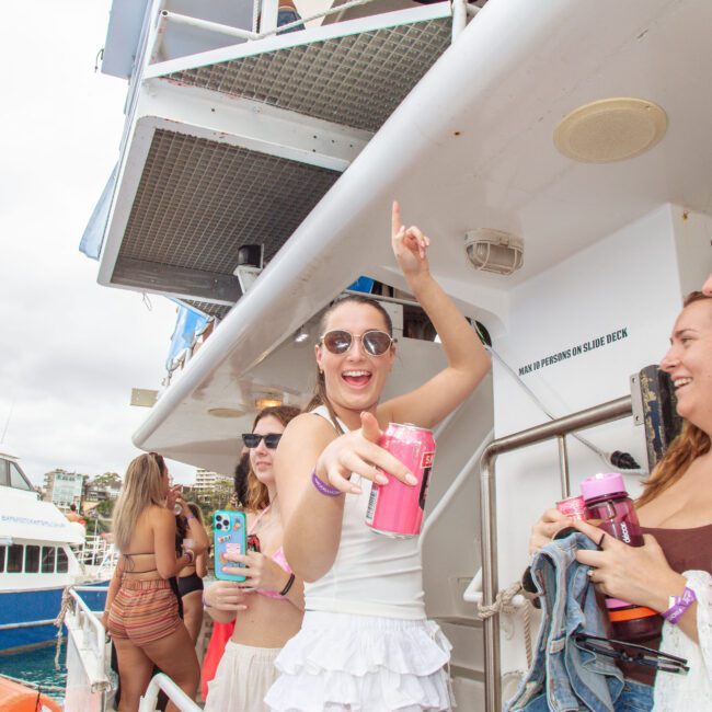 A group of young women in summer clothes stand on a boat, smiling and laughing. One woman in sunglasses points upward while holding a drink. Other boats and umbrellas are visible in the background.